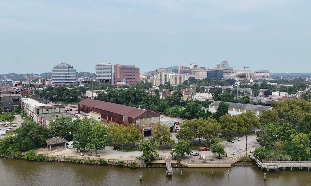 A tree-lined riverbank with brick buildings and a distant Delaware skyline.