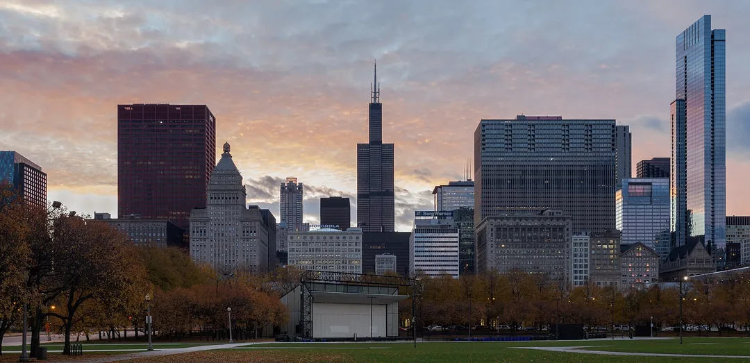 A tall, dark skyscraper rises above the Illinois skyline beyond an open park.