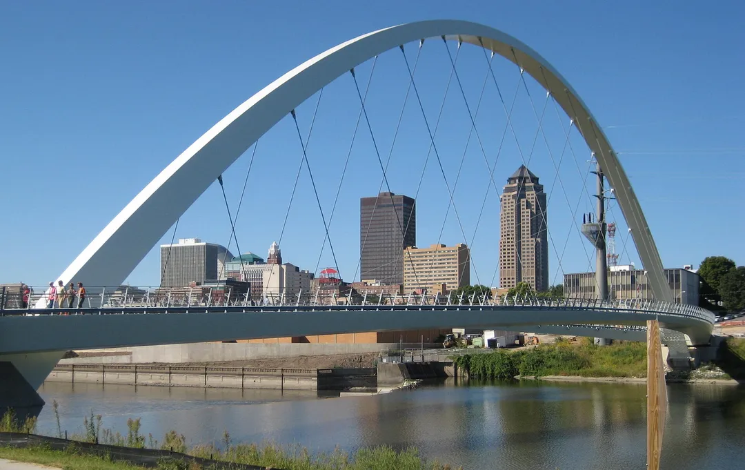 A white arched pedestrian bridge frames downtown buildings across an Iowa river.