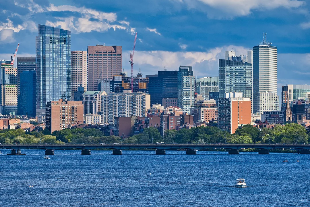 A Massachusetts skyline rises beyond a tree-lined riverbank and blue water.
