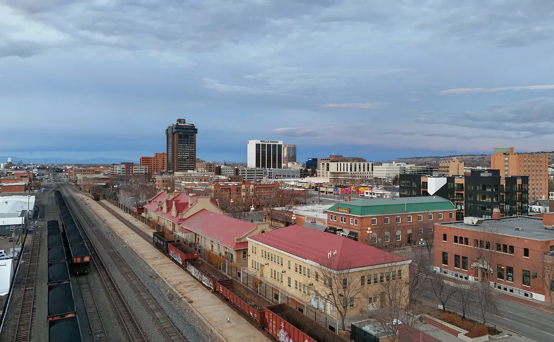 Rail lines and freight cars lead past red-roofed buildings toward downtown towers in Montana.