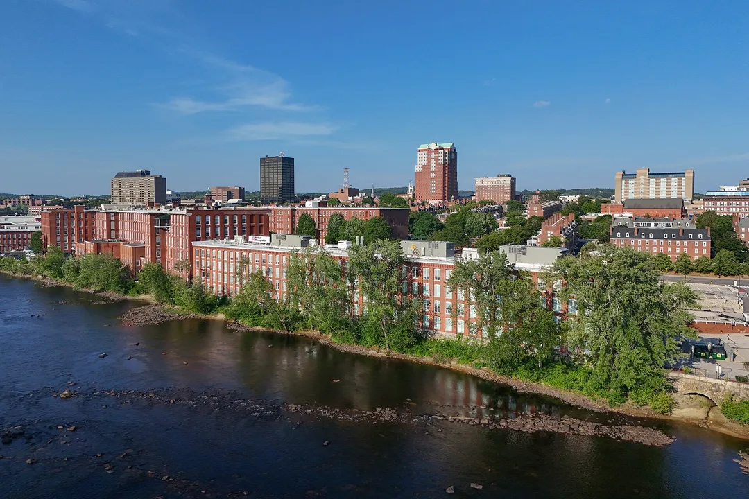 Long brick buildings line a tree-covered riverbank beneath New Hampshire city towers.