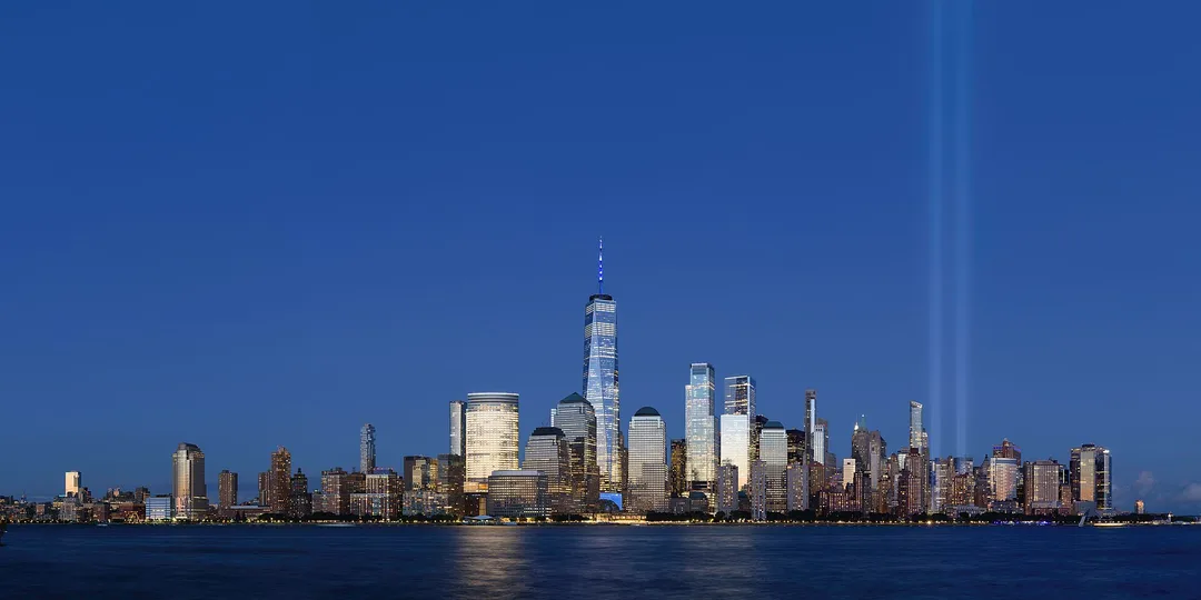 Manhattan's illuminated waterfront skyline across the water at dusk, with two beams of light rising into the sky.