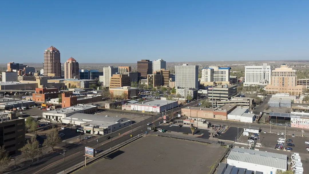 Downtown towers rise above low buildings, roads, and parking lots in a broad New Mexico cityscape.