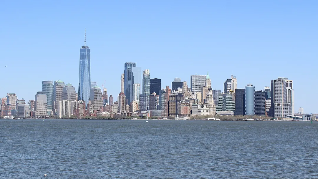 Manhattan skyscrapers line the waterfront beyond rippling water under a clear blue sky.