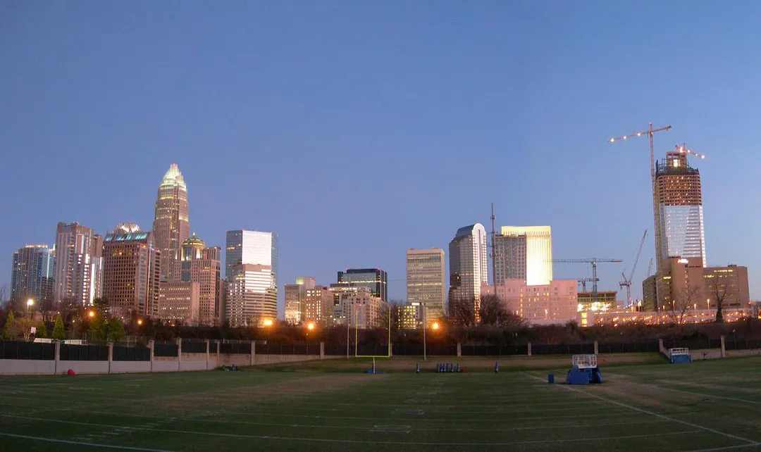 Lighted office towers and a construction crane stand beyond a grassy sports field at dusk in North Carolina.