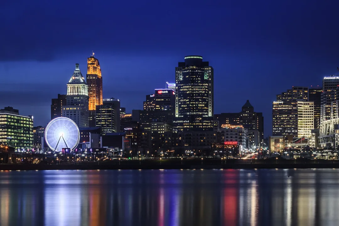 A glowing Ferris wheel and Ohio city towers cast colorful reflections across a river at night.