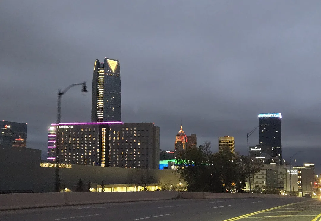 An illuminated glass tower rises above an Oklahoma street beneath a gray evening sky.