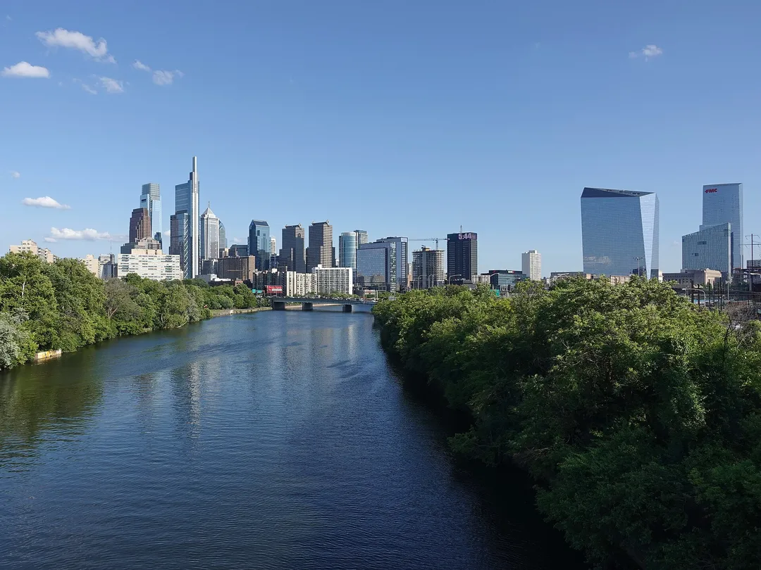 A river bordered by dense green trees leads toward Pennsylvania skyscrapers beneath scattered clouds.
