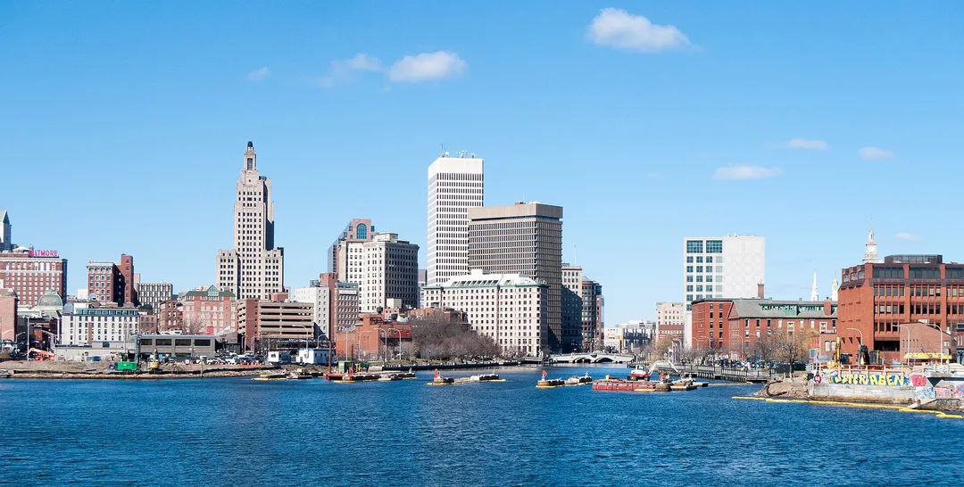 Small boats sit along a Rhode Island waterfront backed by brick buildings and tall office towers.