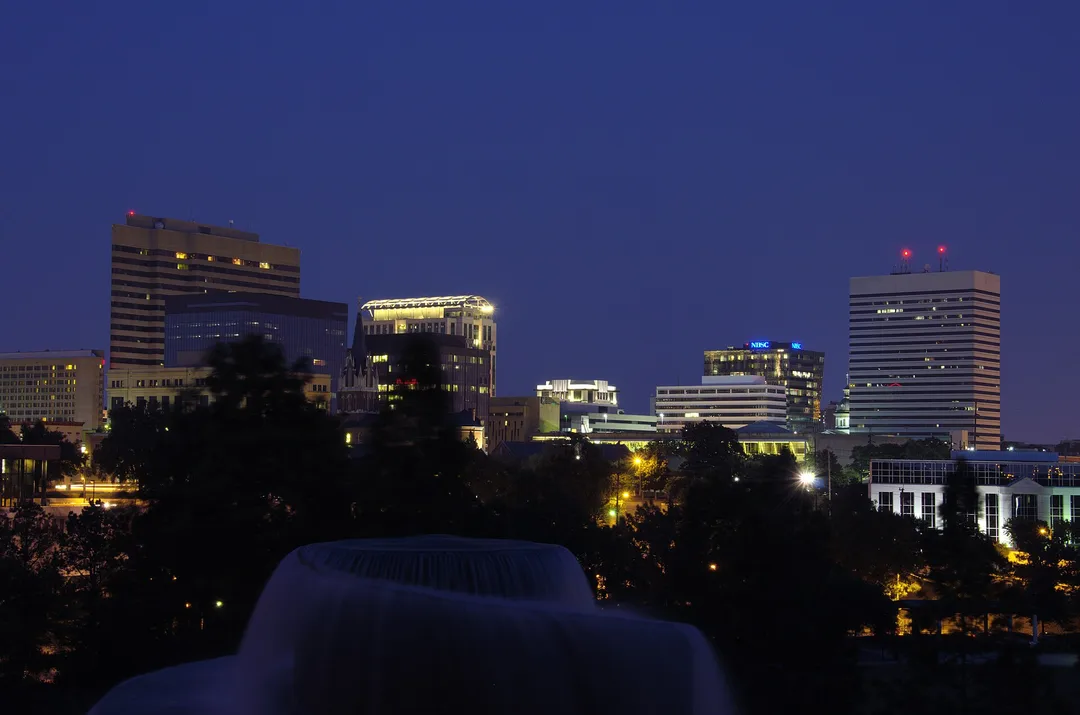 Lighted office buildings rise above dark trees and a fountain at night in South Carolina.