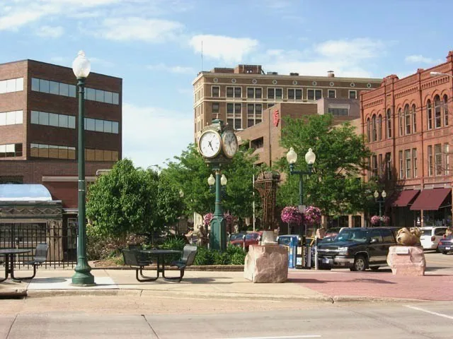 A street clock and hanging flower baskets stand beside brick storefronts in South Dakota.