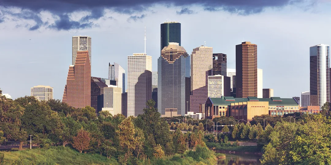 A cluster of Texas skyscrapers rises above a broad canopy of green trees beneath drifting clouds.