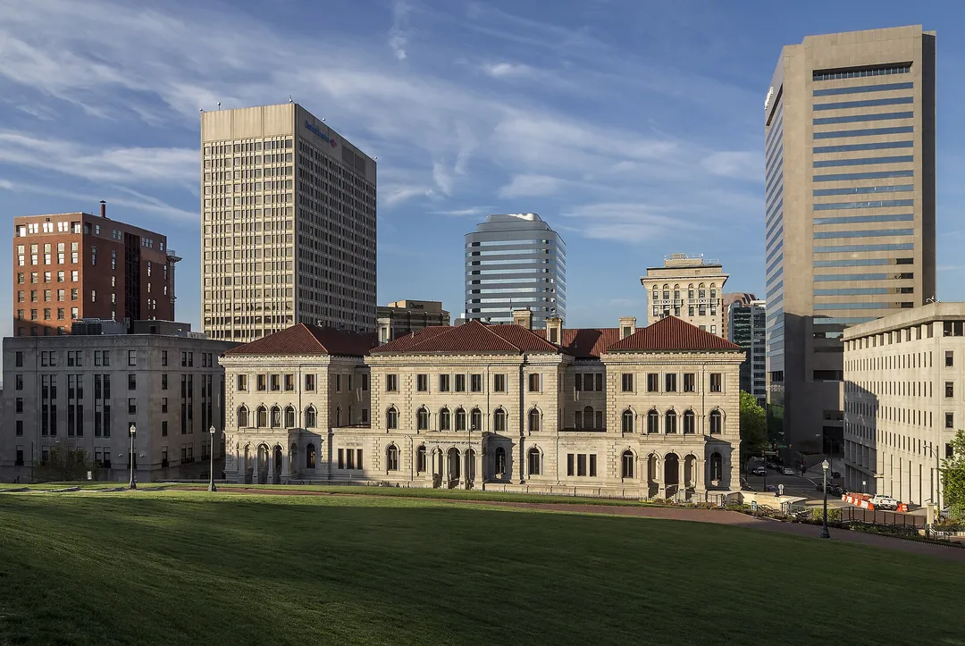 An ornate stone building faces a wide lawn with modern Virginia office towers rising behind it.