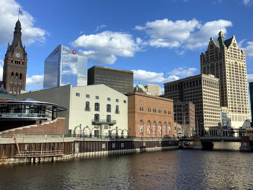 Historic riverfront buildings, a clock tower, and modern high-rises line a Wisconsin waterway.